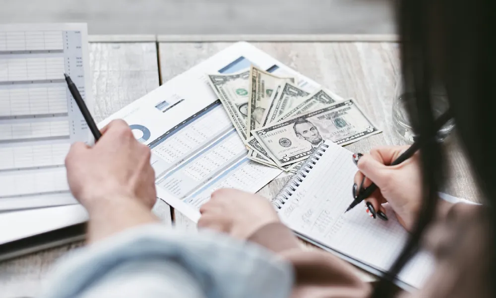 Dos personas analizando gráficos financieros, billetes de dólar y tomando notas en una libreta sobre una mesa de madera, representando el cálculo del costo de un seguro de vida.