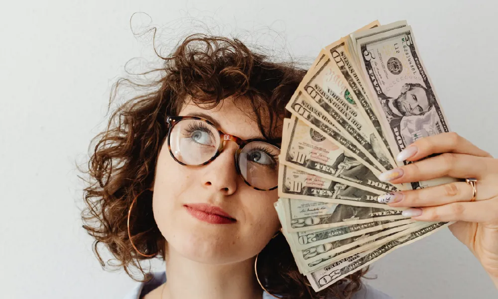 Mujer con gafas y cabello rizado mirando pensativamente hacia arriba, sosteniendo un fajo de dólares en la mano, representando el cálculo del costo de un seguro de vida.