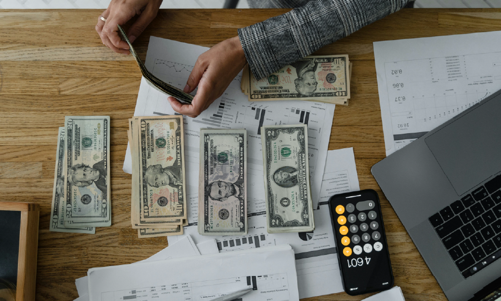 Vista cenital de una persona contando fajos de billetes sobre un escritorio con documentos financieros, una calculadora en un celular y una laptop.
