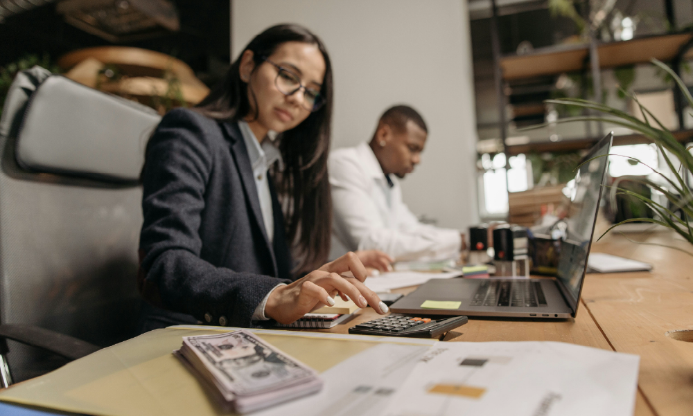 Una profesional de finanzas con lentes usa una calculadora en su escritorio de oficina, con un fajo de billetes y una laptop. Un colega trabaja en el fondo.