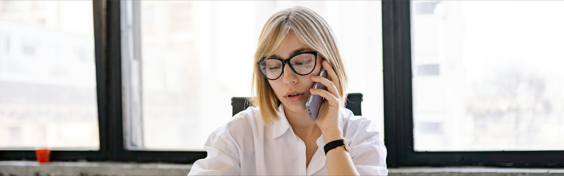 Una mujer joven con gafas, vestida con una camisa blanca, sentada en un escritorio frente a una ventana, hablando por teléfono móvil mientras escribe notas en un portapapeles.