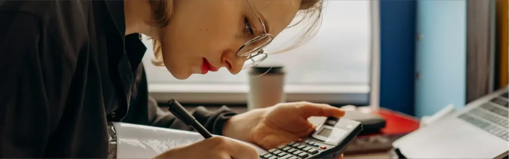 Mujer joven en oficina con gafas escribiendo en un cuaderno y usando una calculadora, analizando opciones de seguro de vida antes de los 30.