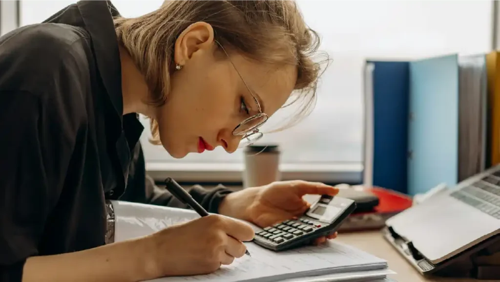 Mujer joven en oficina con gafas escribiendo en un cuaderno y usando una calculadora, analizando opciones de seguro de vida antes de los 30.
