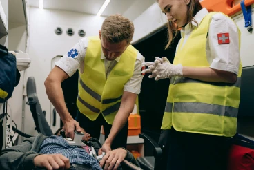 Primer plano dramático dentro de una ambulancia: un paramédico usa un desfibrilador en el pecho de un hombre que yace inconsciente, mientras una colega vestida con uniforme y guantes observa. Representa una emergencia médica grave que puede ser cubierta por seguros de vida.