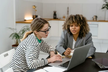 Dos mujeres profesionales de seguros, una con gafas y otra con pelo rizado, colaborando frente a una laptop y tomando notas en una oficina moderna.