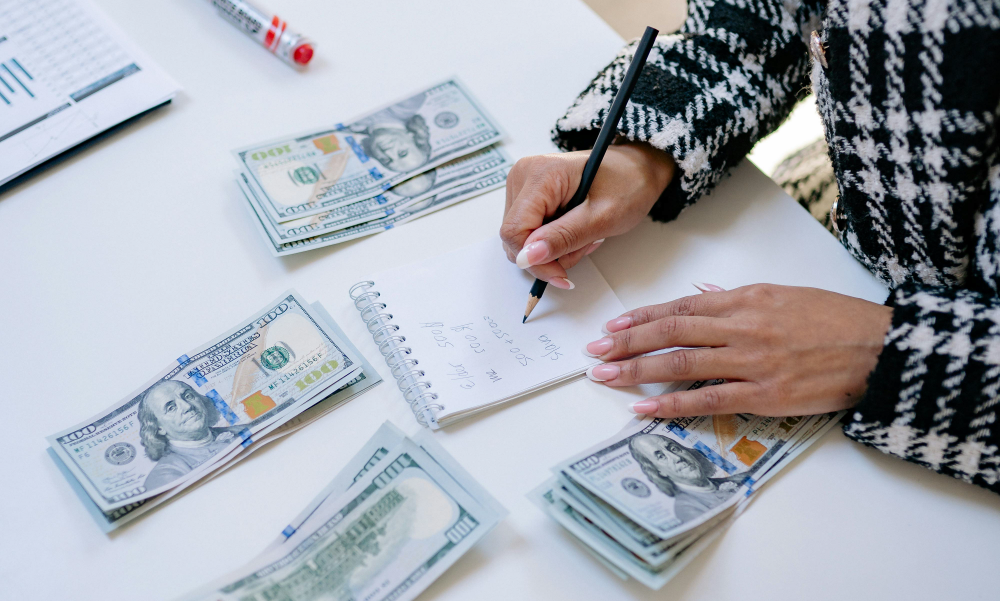 Primer plano de manos femeninas escribiendo cálculos de ahorro en un cuaderno junto a varios montones de billetes de 100 dólares.