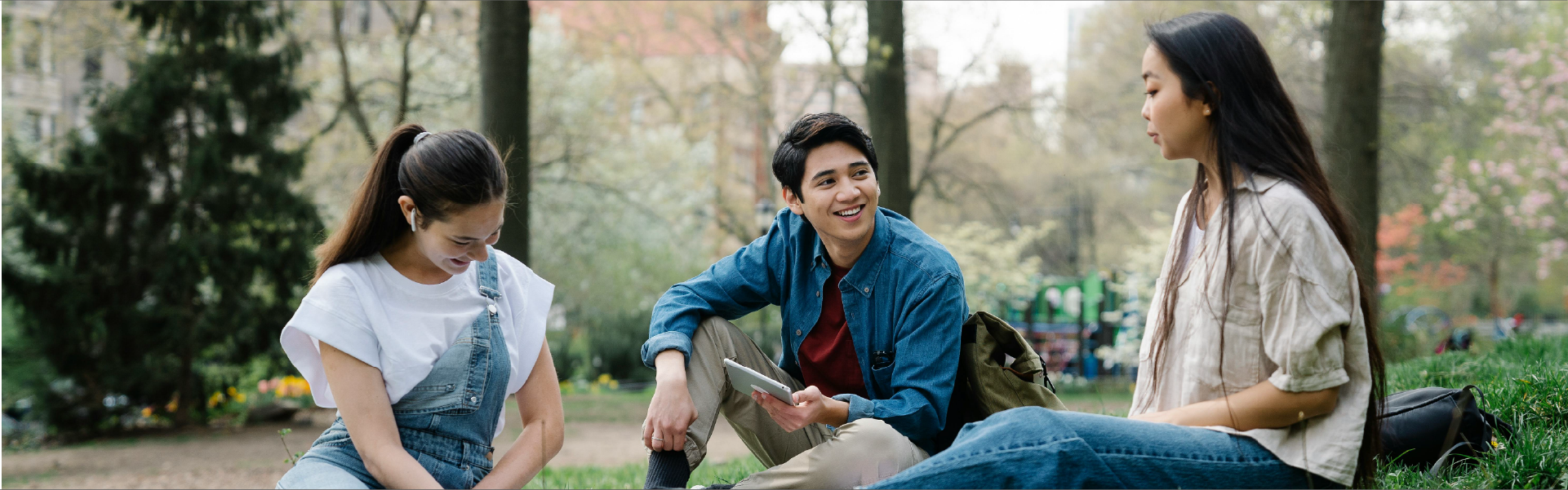 Tres jóvenes universitarios sentados en el césped de un parque al aire libre. A la izquierda, una joven con overol de jean y camiseta blanca mira hacia abajo sonriendo; en el centro, un joven con camisa de jean abierta sostiene una tableta digital mientras conversa animadamente; a la derecha, otra joven de cabello largo escucha la conversación de perfil. Al fondo se aprecian árboles y naturaleza difuminada.