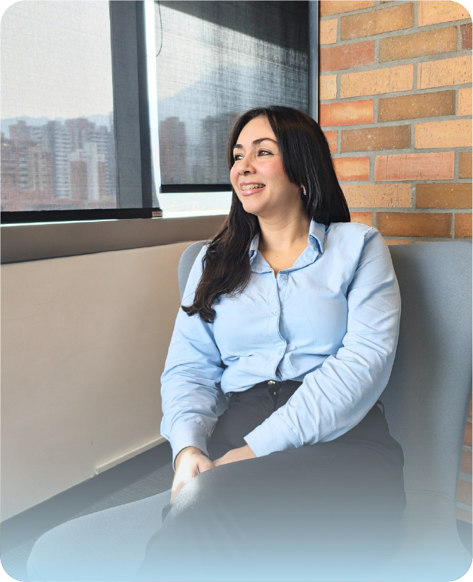 Especialista de la agencia Visión Segura sonriendo sentada en un sofá gris, mirando hacia un lado. Viste una camisa formal azul claro, pantalón negro y se encuentra en una oficina moderna frente a un ventanal y una pared de ladrillos expuestos.
