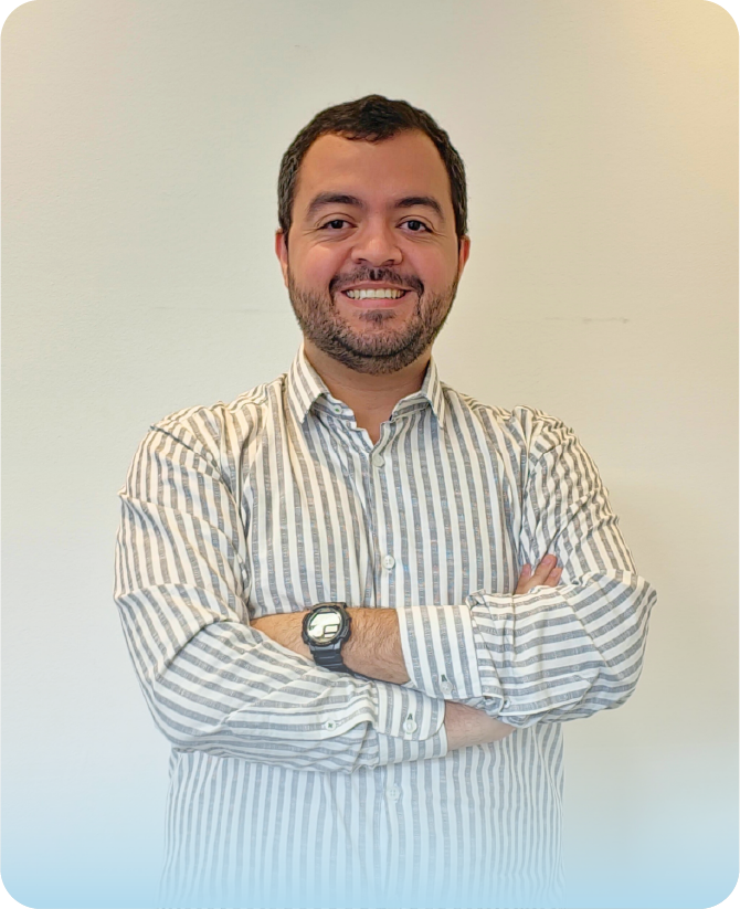 Especialista de la agencia Visión Segura sonriendo con los brazos cruzados frente a un fondo blanco. Viste una camisa formal a rayas verticales claras y un reloj deportivo negro en su muñeca.