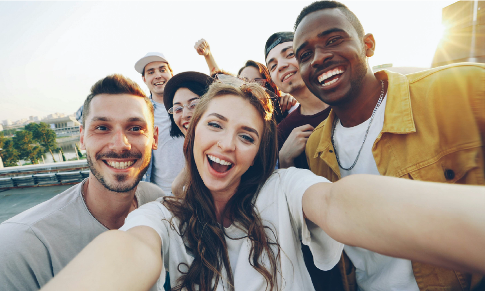Grupo de amigos jóvenes de diversas etnias sonriendo con entusiasmo mientras se toman una foto estilo selfi al aire libre en una terraza durante el atardecer. En primer plano destaca una joven de cabello castaño claro sonriendo con los brazos extendidos sosteniendo la cámara, acompañada a los lados por varios amigos alegres.