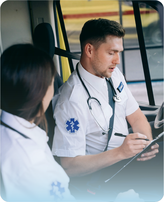 Dos paramédicos o personal de asistencia médica dentro de una ambulancia. Un enfermero con uniforme blanco y estetoscopio al cuello escribe notas en una tabla portapapeles, mientras una compañera en primer plano lo acompaña.