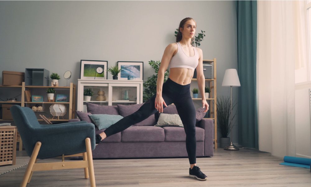 Mujer realizando estiramientos y ejercicios de pierna apoyada en una silla en su sala, representando la prevención, autonomía y bienestar físico del Programa Longevo de SURA que viene como beneficio del seguro de vida.