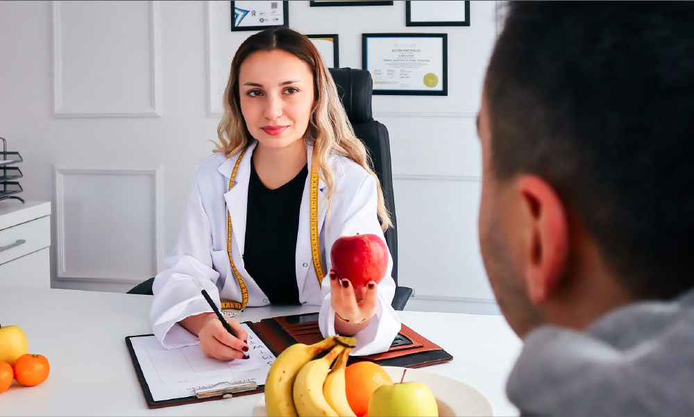 Una nutricionista con bata blanca y cinta métrica en el cuello ofrece una manzana roja a un paciente durante una consulta médica de bienestar y alimentación saludable.
