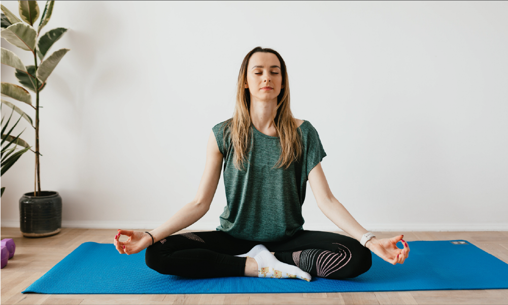 Mujer sentada en postura de loto meditando sobre un mat azul en su casa, representando la salud mental y el bienestar integral cubiertos por un seguro de salud.