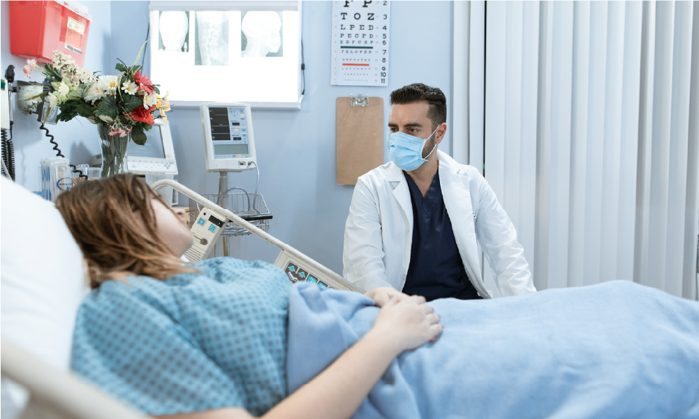 Médico con tapabocas atendiendo a paciente hospitalizada en habitación clínica para ilustrar la cobertura de medicina prepagada o póliza de salud en Colombia.