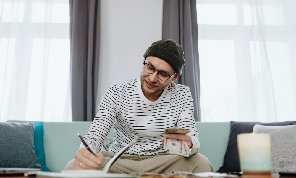 Hombre joven colombiano organizando sus finanzas personales y planeando sus aportes a pensión y seguro de vida, sentado en un sofá con dinero en efectivo y un cuaderno.