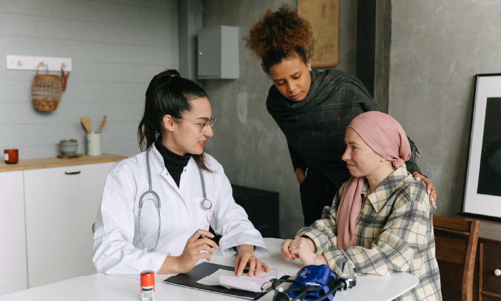 Doctora médica explicando requisitos e informes médicos a una paciente con pañuelo rosa y a su acompañante para reclamar el seguro de vida con cobertura de cáncer.