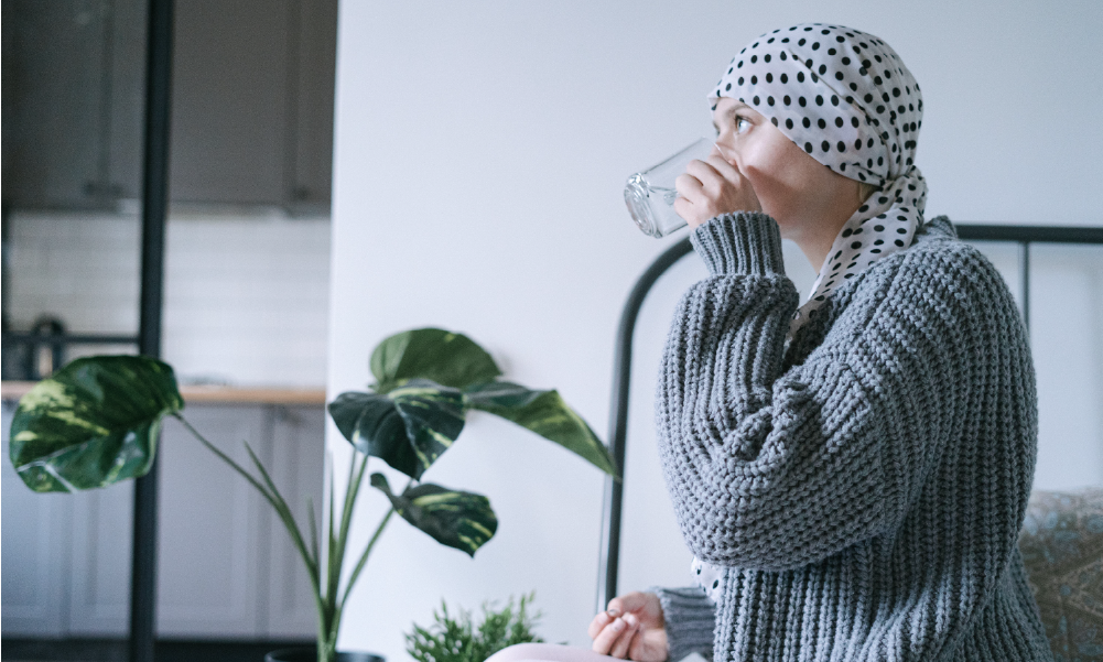 Mujer con pañuelo en la cabeza tomando agua y medicamentos, representando la cobertura y apoyo de un seguro de vida con cobertura de cáncer.